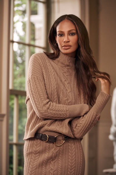 Elegant woman wearing a beige cable-knit sweater and matching skirt, standing indoors near large windows, showcasing professional fashion photography for stylish and natural portrait images.