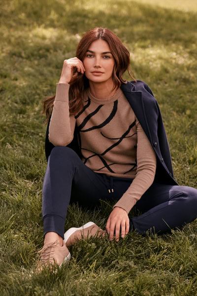 Casual fashion portrait of a young woman sitting on grass, wearing a beige sweater with black abstract lines, navy pants, and a navy jacket, under natural sunlight, outdoors, in a lifestyle and fashion photography style.