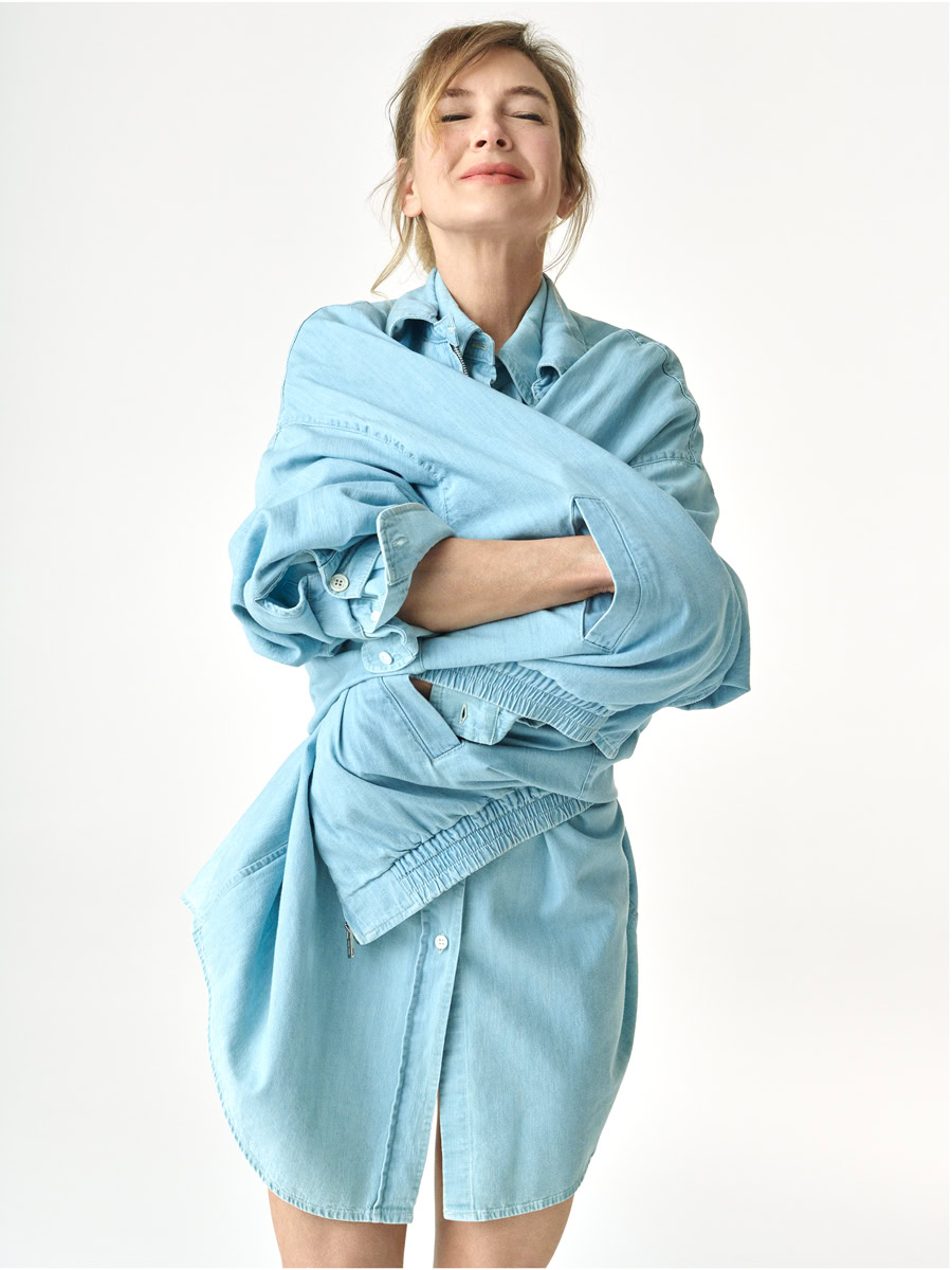 Bright young woman in a light blue oversized denim jacket and matching skirt, posing with arms crossed and a confident smile, showcasing modern fashion photography by Pamela Hanson.