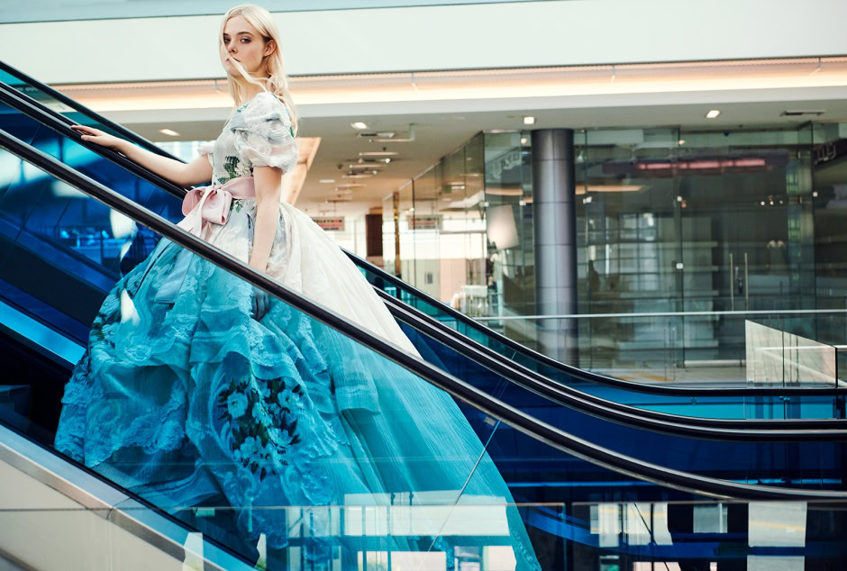 Elegant woman in a floral and pastel gown ascending an escalator in a modern shopping mall, showcasing high fashion photography by Pamela Hanson.
