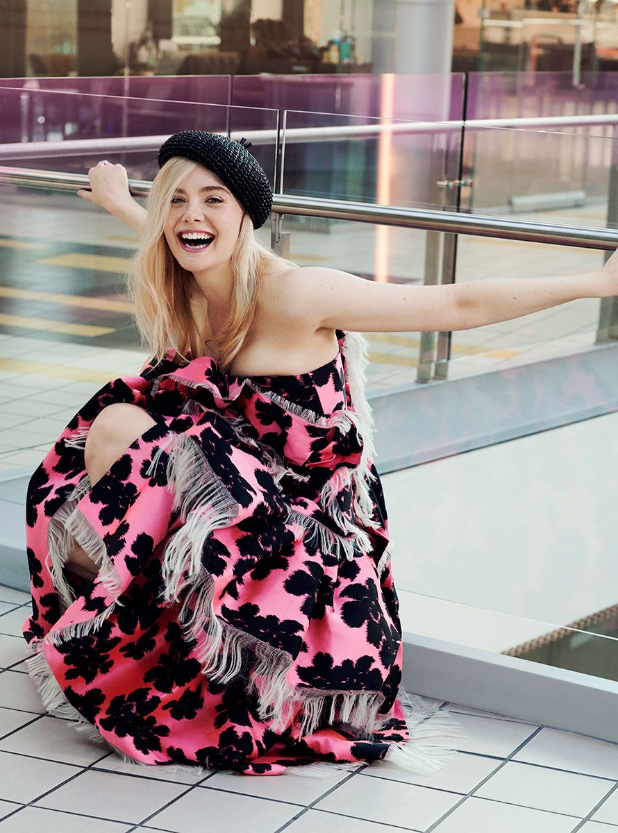 Colorful fashion portrait of a young blonde woman smiling, wearing a black beret and a bold pink and black floral dress, in a modern urban setting with glass railing and contemporary architecture.