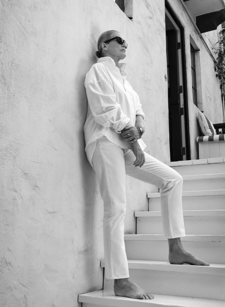 Black and white portrait of mature woman in stylish white outfit with sunglasses posing on stairs at modern home exuding elegance and confidence.