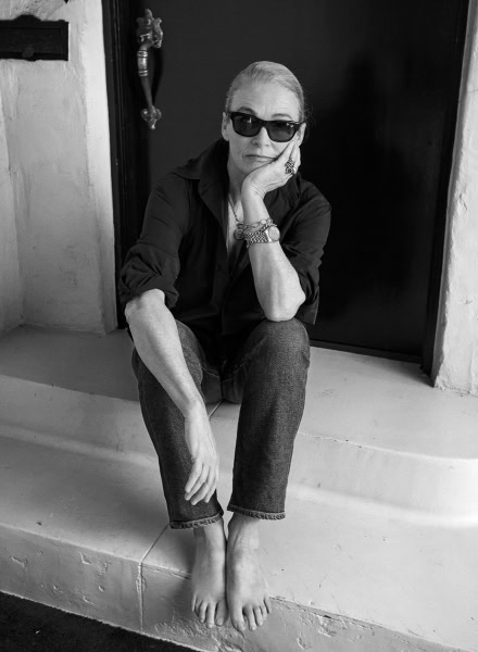 Elegant woman wearing sunglasses sitting barefoot on a doorstep in a black and white fashion portrait.