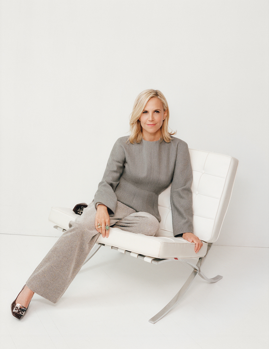 Elegant blonde woman in stylish gray sweater and checkered pants sitting on modern white leather lounge chair in minimalist studio setting.