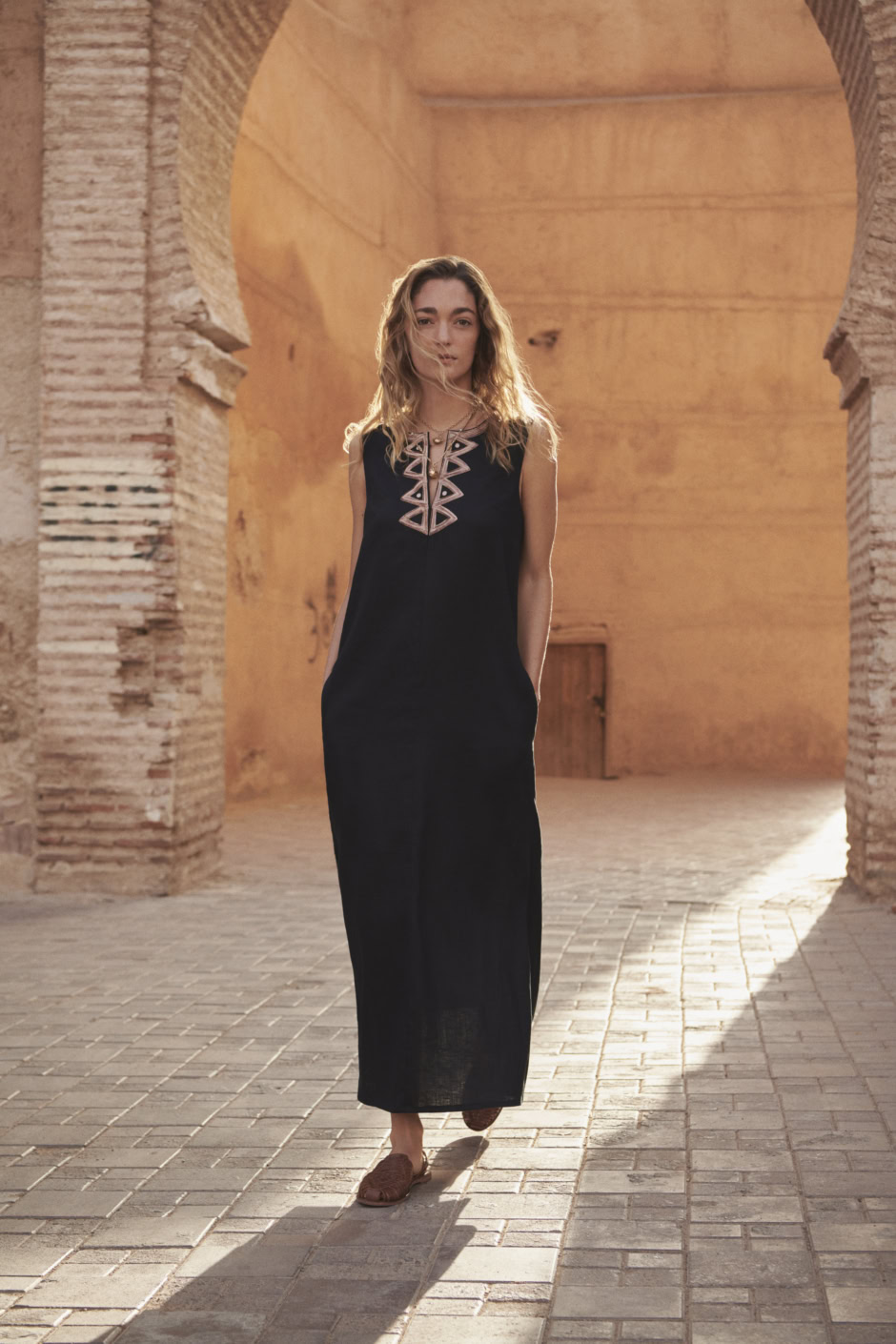 A young woman in a long black dress with embroidered top standing outdoors amid ancient brick arches, capturing elegant fashion photography in warm sunset light.