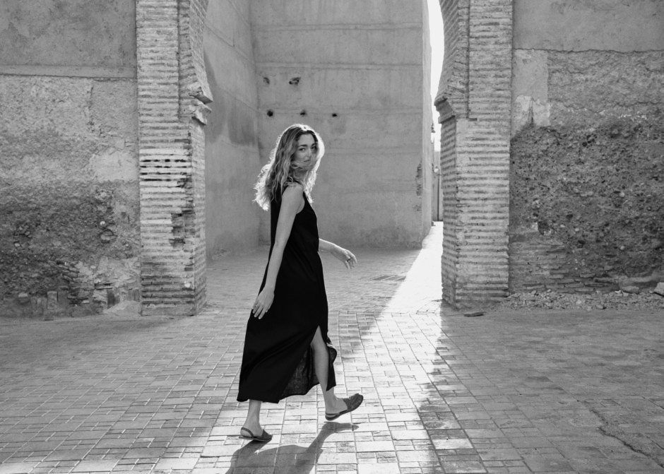 Elegant woman walking through historic stone archway, black dress and sandals, illuminated by natural sunlight, capturing artistic fashion photography.