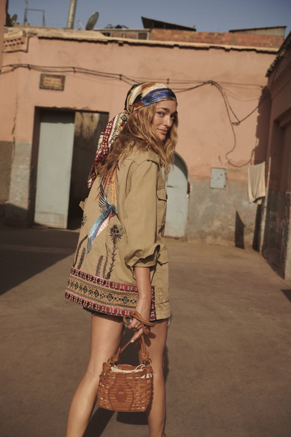 Colorful bohemian woman with headscarf holding wicker handbag in urban street scene.