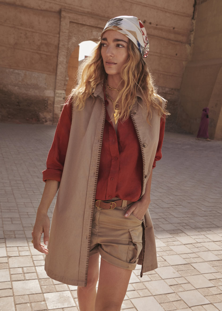Elegant woman wearing a red blouse, beige vest, and shorts with a colorful headscarf, captured in a warm, sunlit outdoor setting with historic architecture in the background.