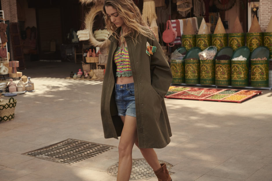 Colorful boho street style woman wearing a patchwork crop top, oversized jacket, denim shorts, and boots walking through a vibrant marketplace.