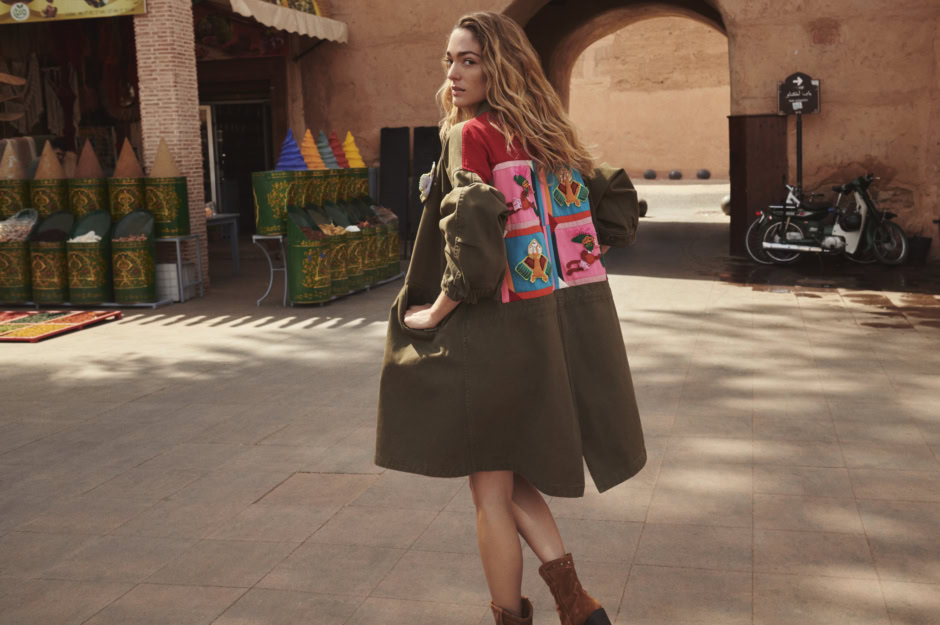 Vibrant fashion photography featuring a woman wearing a stylish, colorful patchwork jacket and brown boots in an outdoor market setting; fine art portrait by Pamela Hanson.