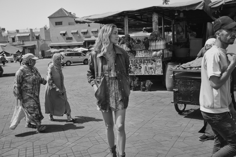 Vintage street photography of a young woman walking in a market, capturing candid moments of daily life in a bustling urban setting with diverse pedestrians and colorful vendor stalls.