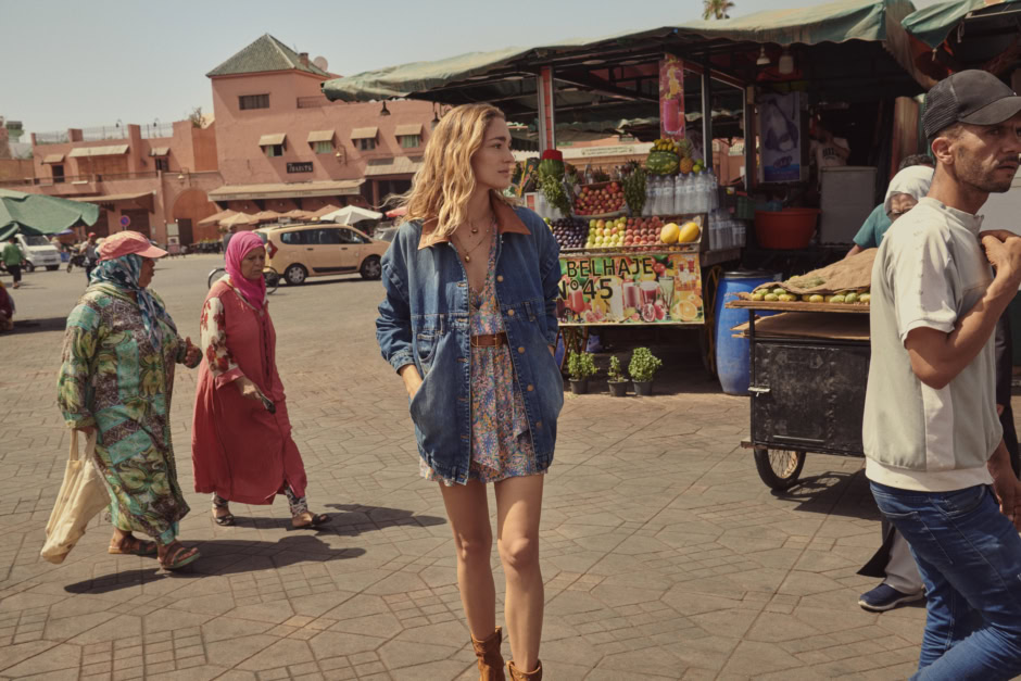 Vibrant street scene featuring a stylish woman in a colorful dress and denim jacket walking through an outdoor market with local vendors and traditional architecture in the background, capturing authentic Moroccan culture.