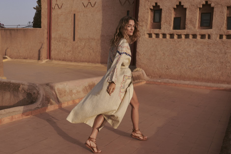 Elegant woman in bohemian styled dress and sandals walking outdoors at sunset, adobe-inspired architecture, warm tones, desert landscape, stylish fashion photography, Pamela Hanson Photography.