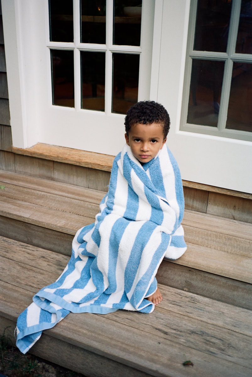 Boy wrapped in blue and white striped towel sitting on wooden steps outside house, candid portrait, natural light, urban lifestyle photography.