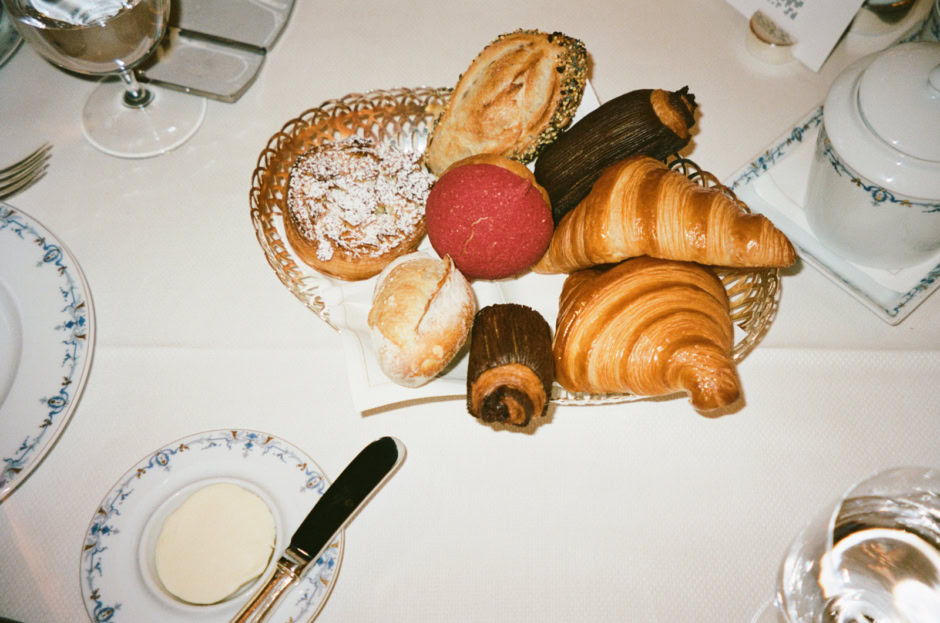 Freshly baked croissants, bread rolls, and assorted pastries in a basket, served with butter and beverages, captured in a professional food photography style by Pamela Hanson.