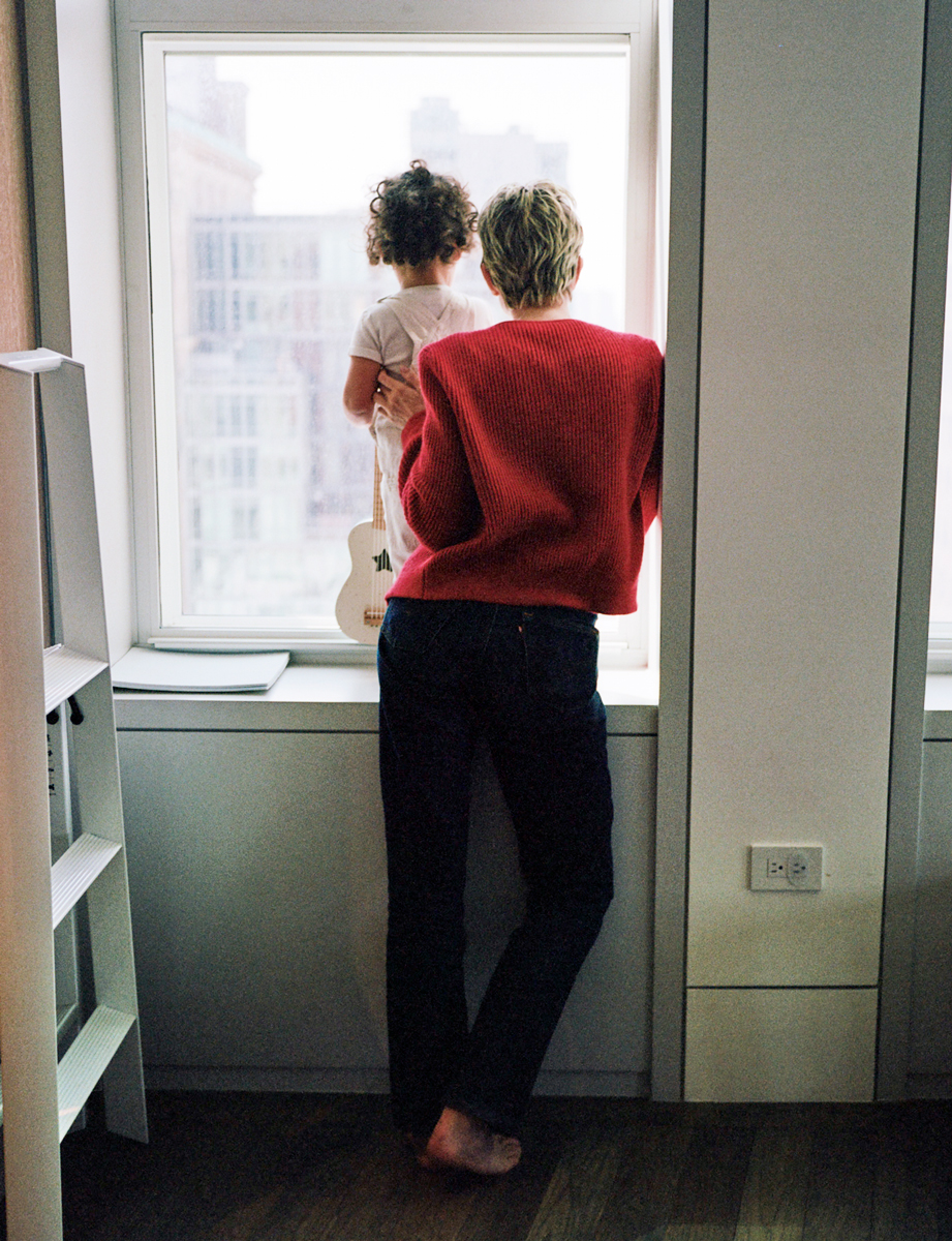 Child looking out the window with an adult, urban cityscape background, cozy indoor setting, contemporary lifestyle photography, Pamela Hanson Photography.