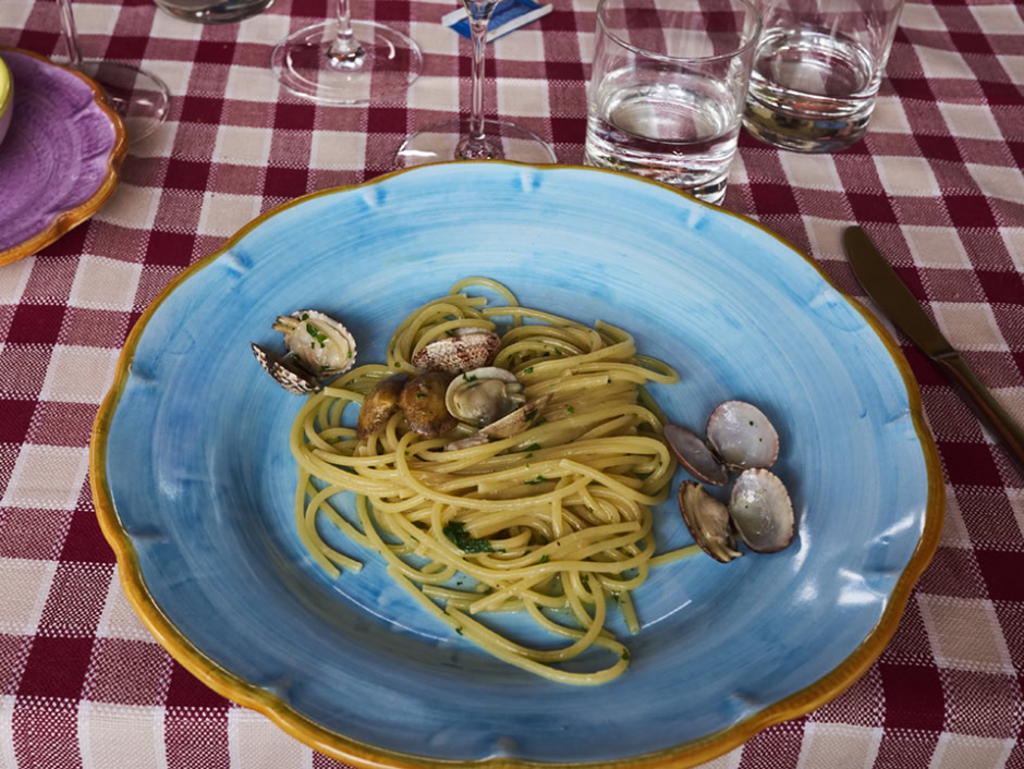 Vibrant blue ceramic plate with linguine pasta and fresh seafood, on a red and white checkered tablecloth, styled with elegant glassware and cutlery, showcasing gourmet Italian cuisine.