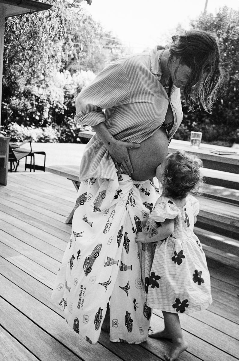 Pregnant woman hugging her young daughter outdoors on a wooden deck, capturing a tender maternity and family moment in black and white.