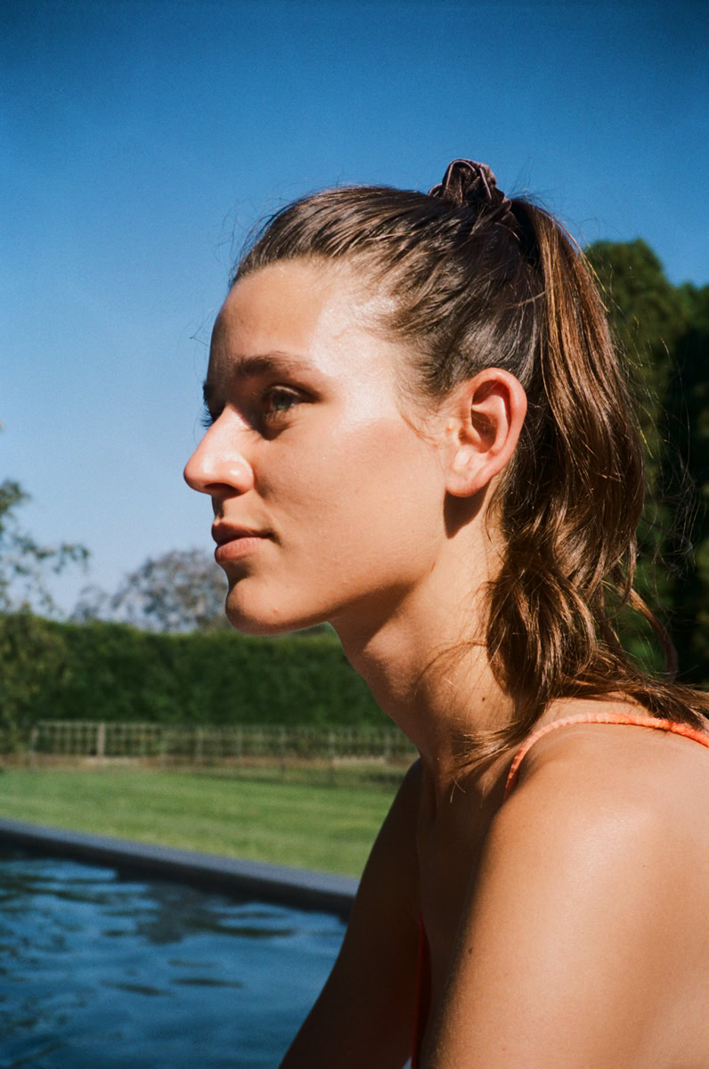 Young woman portrait outdoors by a pool, natural lighting, vibrant summer scenery, relaxed and contemplative mood, professional photography by Pamela Hanson.