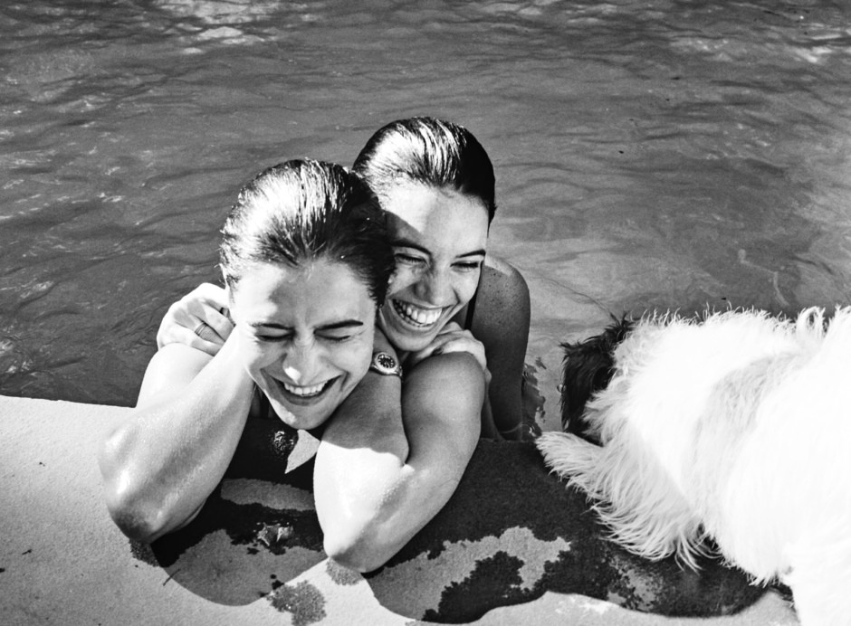 Close-up black and white photograph of two smiling women with wet hair, embracing and enjoying a fun moment at the poolside with their dog.