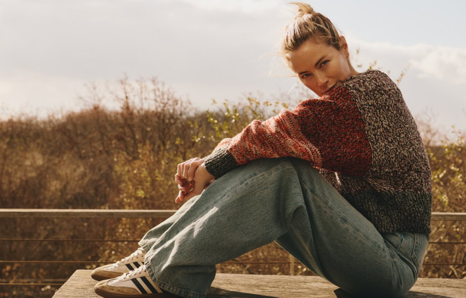 Autumn woman sitting outdoors on a wooden deck, wearing a cozy multicolored knit sweater, jeans, and sneakers, with a natural background of fall foliage and soft sunlight.
