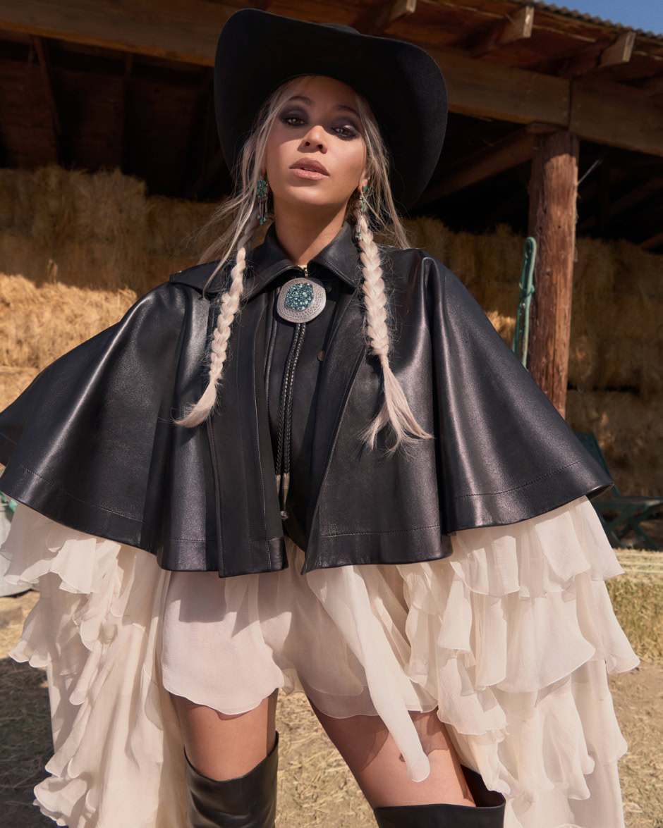 Elegant fashion portrait featuring a model in high-fashion Western-inspired attire, including a wide-brimmed hat, leather cape, and layered ruffled skirt, outdoors in natural sunlight.