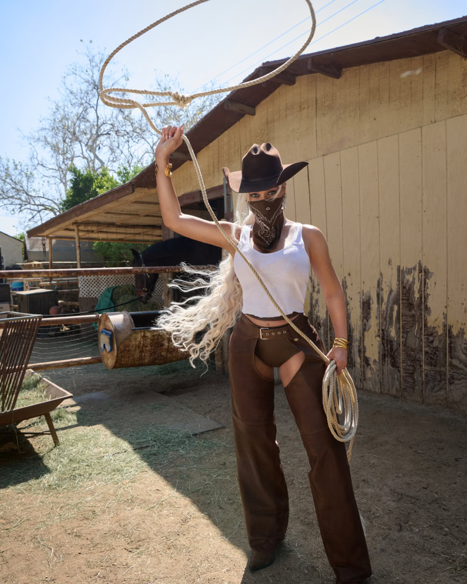 Ranch-themed fashion photoshoot with a woman wearing cowboy hat, bandana, leather pants, and holding a lasso outdoors at a rustic farm setting.