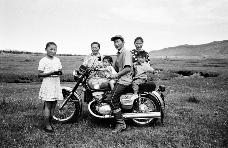 A vintage black-and-white photograph of a family in a rural landscape, featuring a man on a motorcycle with children and women, capturing candid moments, emphasizing traditional life and cultural heritage.