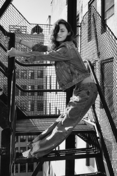 Elegant black and white portrait of a young woman on an urban fire escape, showcasing contemporary fashion photography with a focus on natural beauty and cityscape background.