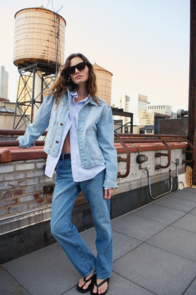 Casual urban rooftop fashion photoshoot featuring a young woman in denim jacket and jeans, showcasing contemporary street style photography on a city rooftop at sunset.