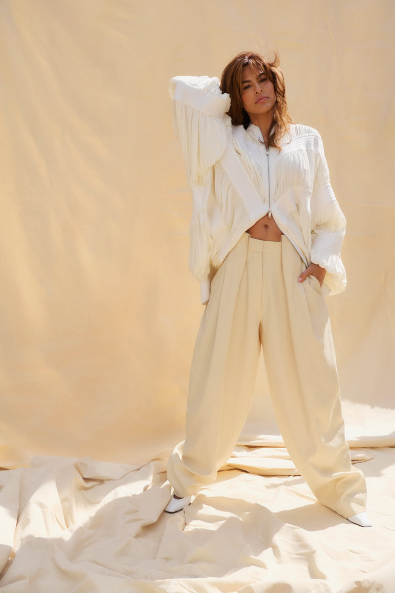Elegant fashion portrait of a woman in cream-colored oversized trousers and a white textured zip-up jacket, standing against a soft beige backdrop. High-end fashion photography by Pamela Hanson.