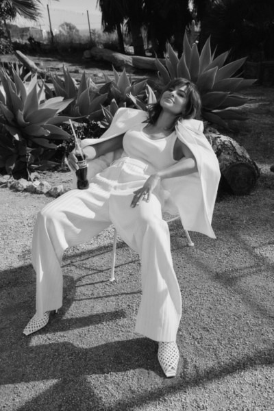Relaxed woman in white outfit sitting outdoors with a Coca-Cola in hand, surrounded by lush plants, black-and-white photography style, showcasing modern fashion photography by Pamela Hanson.