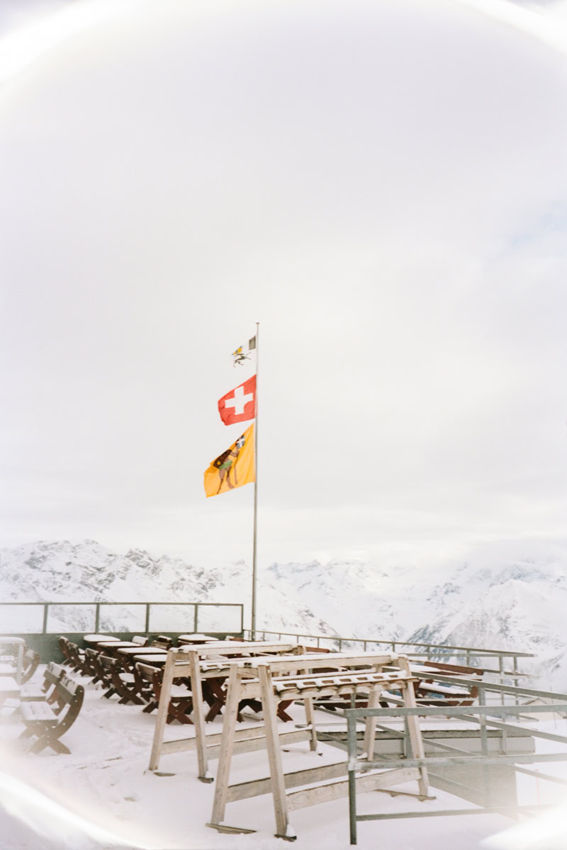 Bright mountain view with Swiss flags, snow-covered terrace chairs, stunning alpine landscape, and cloudy sky, showcasing outdoor architecture photography by Pamela Hanson.