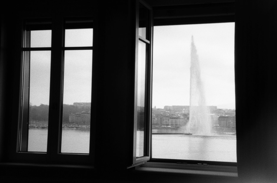 Marvelous black and white photo of a city skyline with a prominent fountain, viewed through a set of open window shutters, capturing urban architecture and water features.