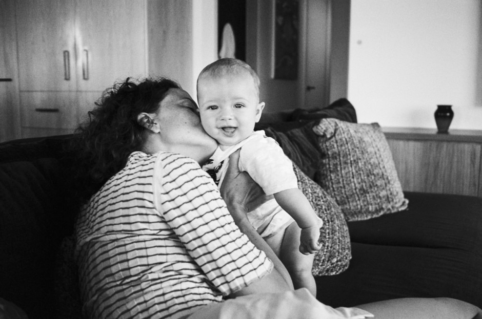 A black and white portrait of a woman holding a smiling baby on a sofa, capturing a heartfelt moment of affection and joy, with a cozy home background.