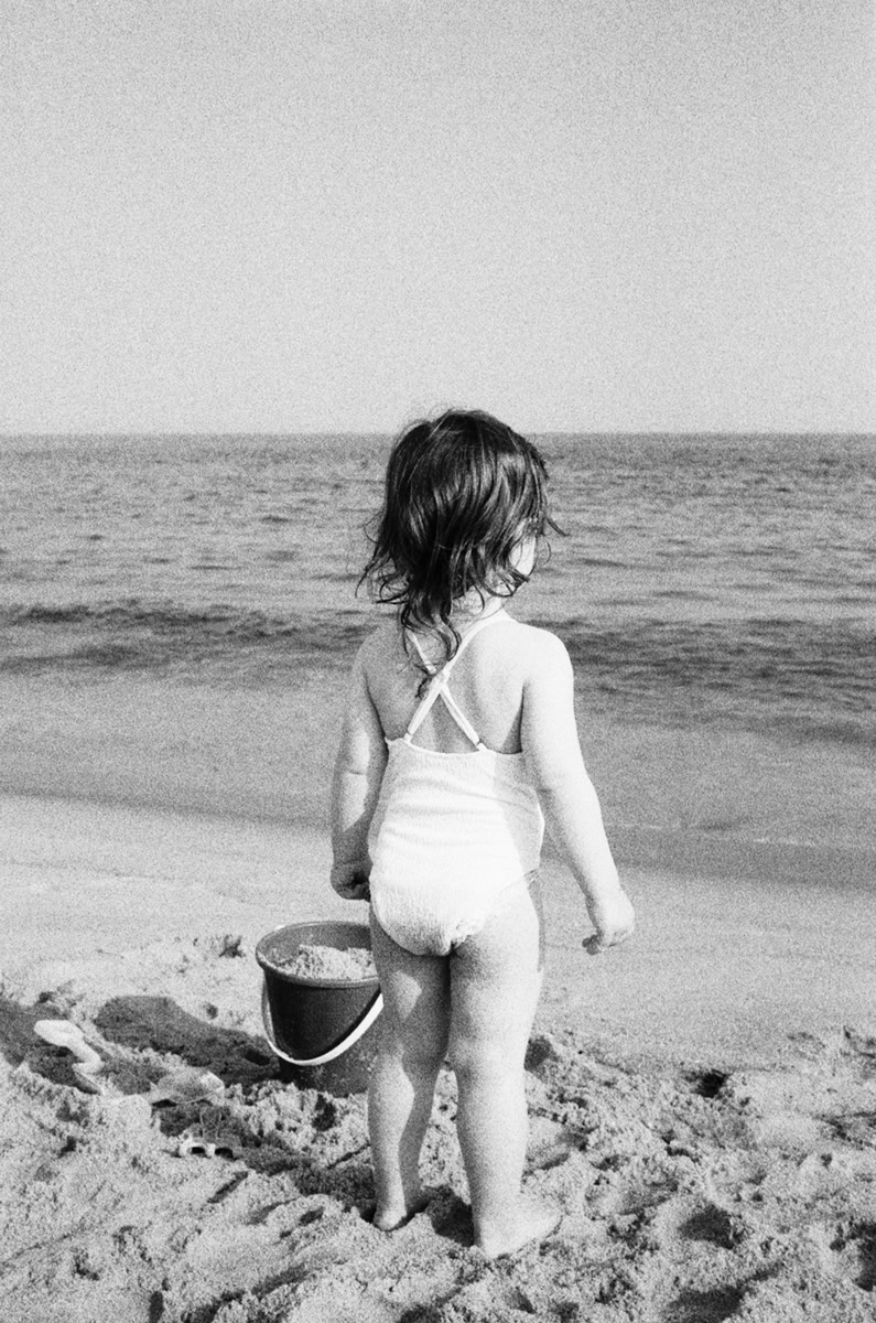 Children at the beach, black and white photo, Pamela Hanson Photography, lifestyle and portrait photography of a young girl with a bucket on a sandy beach, capturing innocence and summertime moments.