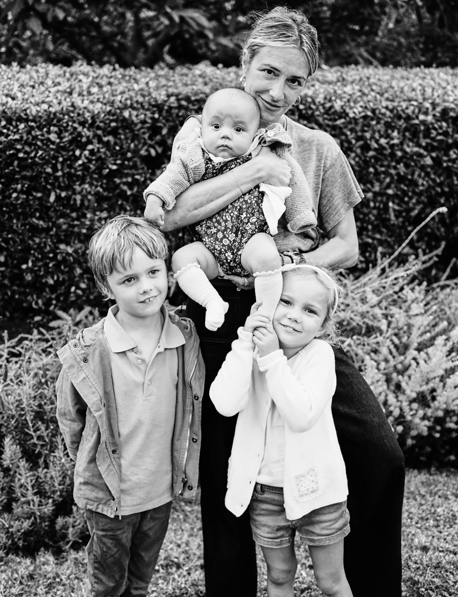 A woman holding two young children outdoors with a garden background in black and white.