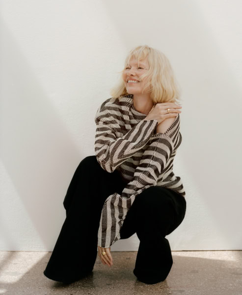 Elegant woman in black pants and checkered sweater smiling, seated against a plain white wall, showcasing portrait photography by Pamela Hanson for fashion and contemporary lifestyle imagery.