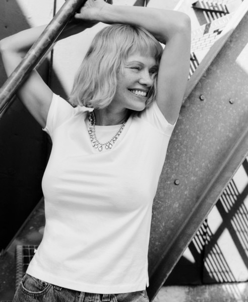 Smiling woman with short blonde hair and layered necklace, casual white t-shirt, outdoor urban setting, black and white photography, contemporary portrait photography, fashion and lifestyle photography.