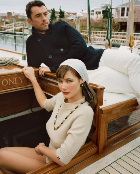Nautical-themed fashion photography featuring a stylish man and woman on a wooden boat dock with boats and houses in the background. Elegant and sophisticated portrait capturing maritime lifestyle.