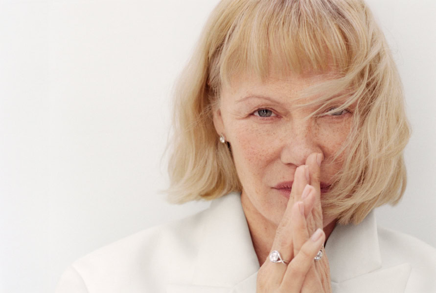 Elegant portrait of a mature woman with blonde hair and blue eyes, posing with her hand near her face against a white background, showcasing professional fashion and beauty photography.