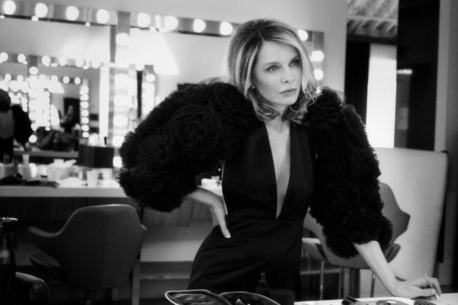Elegant black and white portrait of a woman with blonde hair in a stylish dress with dramatic puffed sleeves, standing at a makeup station in a professional photo studio.