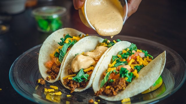 3 tacos on a plate with someone pouring a sauce overtop