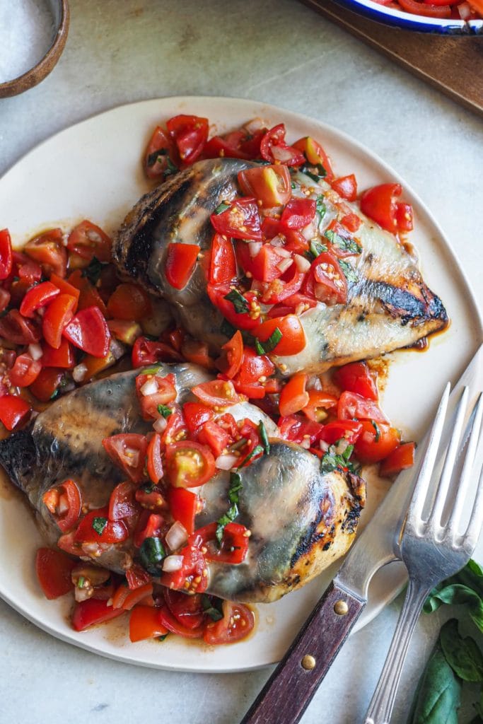 Esta receta de Pechugas de Pollo Caprese es una deliciosa variación de la clásica ensalada italiana! Primero se marina el pollo con un adobo balsámico, para que quede más tierno. Luego se asa a la parrilla, donde el vinagre balsámico se carameliza. Finalmente, se cubre con queso mozzarella y una deliciosa ensalada de tomates cherry con albahaca. Aquí se puede ver en grande un plato con dos pechugas de pollo cocinadas, la mozzarella y la ensalada de tomate por encima. A bajo a la derecha hay un tenedor y un cuchillo apoyados al plato. Arriba a la izquierda hay un pequeño bol con sal.