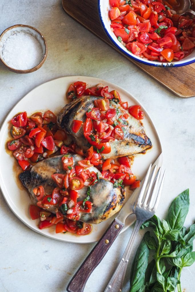 Esta receta de Pechugas de Pollo Caprese es una deliciosa variación de la clásica ensalada italiana! Primero se marina el pollo con un adobo balsámico, para que quede más tierno. Luego se asa a la parrilla, donde el vinagre balsámico se carameliza. Finalmente, se cubre con queso mozzarella y una deliciosa ensalada de tomates cherry con albahaca. Aquí se puede ver un plato con dos pechugas de pollo cocinadas, la mozzarella y la ensalada de tomate por encima. A bajo a la derecha hay un tenedor y un cuchillo apoyados al plato y un manojo de albahaca. Arriba a la izquierda hay un pequeño bol con sal y arriba a la derecha un bol con extra de ensalada encima de una tabla de madera.