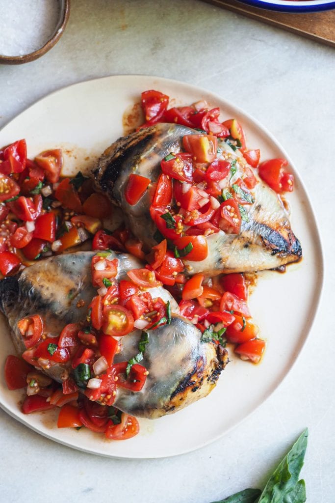 Esta receta de Pechugas de Pollo Caprese es una deliciosa variación de la clásica ensalada italiana! Primero se marina el pollo con un adobo balsámico, para que quede más tierno. Luego se asa a la parrilla, donde el vinagre balsámico se carameliza. Finalmente, se cubre con queso mozzarella y una deliciosa ensalada de tomates cherry con albahaca. Aquí se puede ver un plato con 2 pechugas cocinadas con la mozzarella y la ensalada por encima. Arriba a la izquierda hay un pequeño bol con sal.