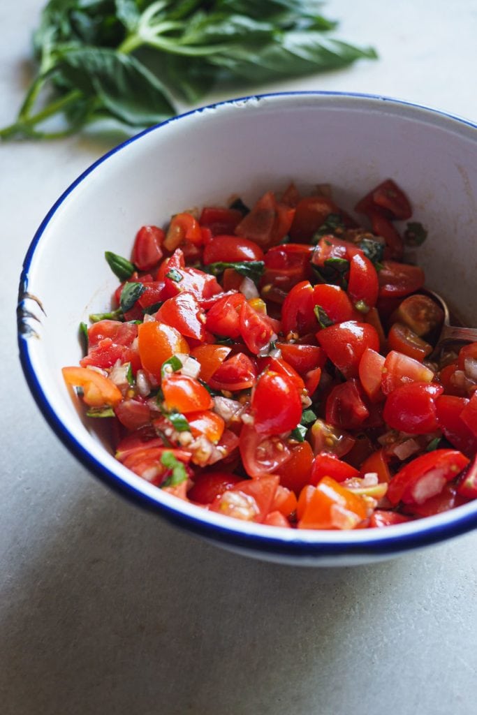 La ensalada caprese se usa como topping para la pechuga de pollo con mozzarella. Hacemos en cantidad para que se pueda servir extra como acompañamiento. Aquí vemos una foto de la ensalada en un bol enamelado con un manojo de albahaca al fondo.
