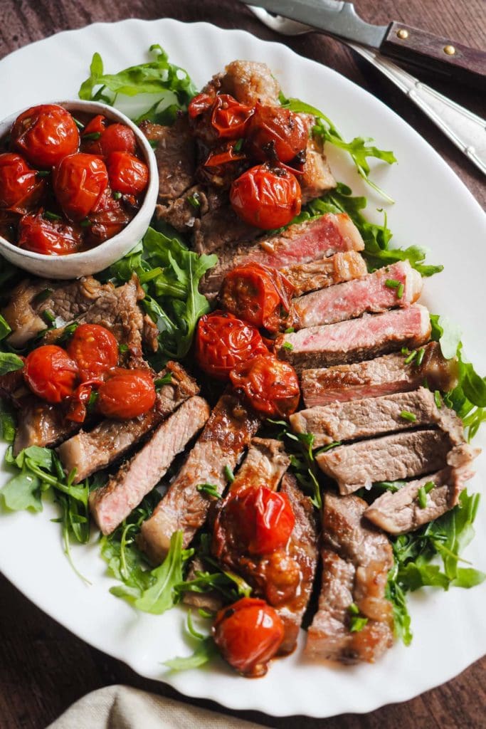 Delicioso Entrecot con Tomates Cherry Asados, Este plato se hace en 15 minutos: mientras la carne reposa, se cocinan los tomates cherry; y si usas una sartén para asar el entrecot, esa misma es la que usarás para prepara toda la comida. Te imaginas el sabor que cogen los tomates? Aquí se ve el corte del entrecot al punto sobre una cama de rúcula y decorado con los tomates cherry asados. Todo servido en una bandeja blanca.