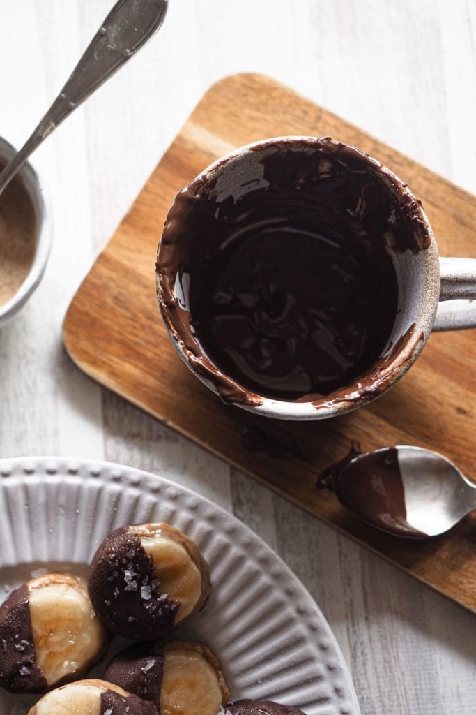 Estos deliciosos Bocaditos Helados de Crema de Almendra Plátano y Chocolate, son un snack perfecto. Son unos caprichitos saludables ideales para los días de verano y, ya que son del tamaño de un bocado, podemos considerarlos bajos en calorías (si logras no comerte más de 1 o 2 a la vez). En esta foto se ve una taza con chocolate derretido usado para dipear los bocaditos.