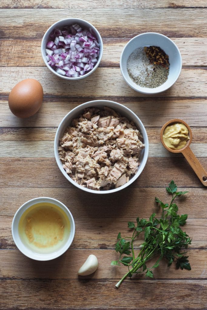 Esta receta de Hamburguesas de Atún es deliciosa, sabrosa y te salva las cenas de entre semana. Están hechas con ingredientes que siempre solemos tener en casa y también son perfectas para preparar comida con antelación! Además, es una receta sin gluten. En esta foto se pueden ver todos los ingredientes: perejil, ajo, atún y su aceite, mostaza, huevo, cebolla morada y especias.